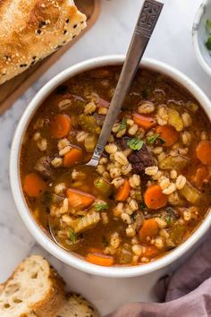 a bowl of soup with carrots, barley and bread on the table next to it