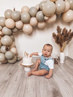 a baby sitting on the floor with a cake and balloons in the air behind him