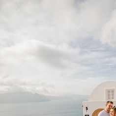 a man and woman standing in front of a white building with the ocean in the background