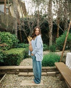 a woman standing in front of a wooden bench and some bushes with trees behind her