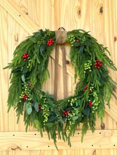 a green wreath with red berries and greenery hangs on a wooden wall next to a fence