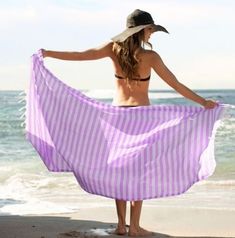 a woman is standing on the beach holding a purple and white striped towel in front of her