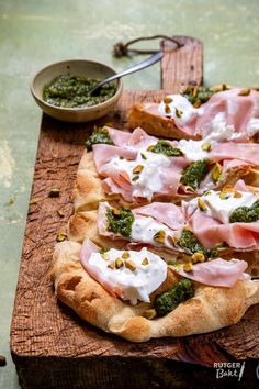 a pizza topped with meat and vegetables on a cutting board next to a bowl of pesto