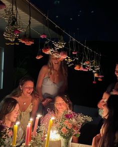 a group of women sitting around a table with candles on it and flowers hanging from the ceiling