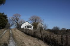 a dirt road next to a fence with a white house on it and trees in the background