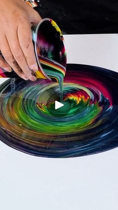 a person pouring paint onto a plate with colorful swirls on it and another hand reaching for the bowl