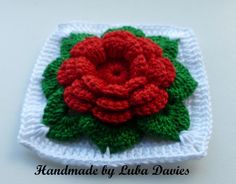 a crocheted red rose on a white square with green leaves in the center