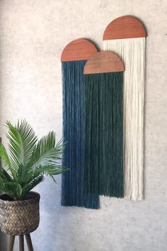 two wall hangings on the side of a white wall next to a potted plant