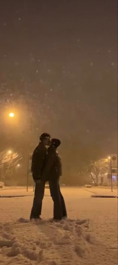 two people standing in the snow at night with their arms around each other and looking into the distance