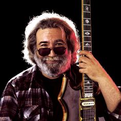 a man with glasses and a beard holding a guitar in his right hand, smiling at the camera