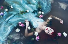 a woman with red hair and blue dress floating in water surrounded by white and pink flowers