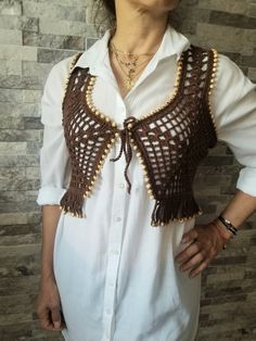 a woman standing in front of a brick wall wearing a white shirt and brown crocheted vest