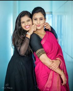 two women in black and pink saris hugging each other with their arms around one another