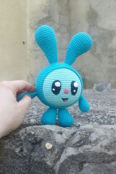 a hand holding a small stuffed animal in front of a stone wall with a building behind it