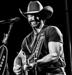 a man wearing a cowboy hat and holding a guitar in front of a microphone on stage