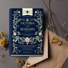 a blue and gold wedding card on top of an envelope next to dried yellow flowers