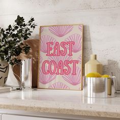 a pink poster with the words east coast on it sitting on a kitchen counter next to a potted plant