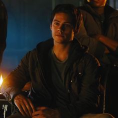 a young man sitting in front of a fire with other people around him looking at the camera