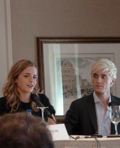 two people sitting at a table with wine glasses in front of them and one person holding a microphone