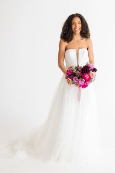 a woman in a white wedding dress holding a pink and purple bouquet with her right hand on her hip