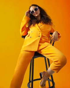 a woman sitting on top of a stool wearing sunglasses and a yellow suit with buttons