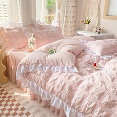 a bed with pink comforters and pillows in a bedroom next to a window filled with white curtains