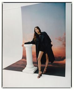 a woman standing next to a tall white column in front of a sky background with clouds