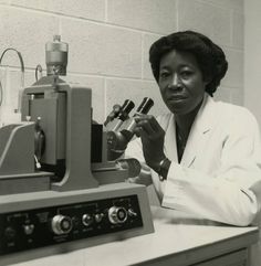 a black and white photo of a woman in a lab coat looking into a microscope