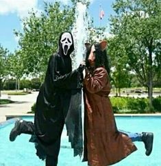 two people dressed in costumes standing next to each other near a fountain with trees and bushes behind them