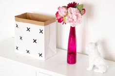 a pink vase with flowers in it next to a small white cat and paper bag