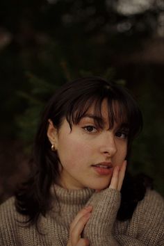 a woman with dark hair wearing a sweater and holding her hand to her face while looking off into the distance