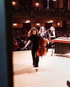 two women are walking down the runway with cellos in their hands and an audience behind them