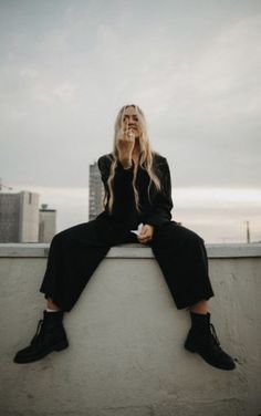 a woman sitting on top of a cement wall talking on a cell phone while wearing all black