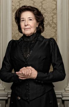 an older woman in a black dress standing with her hands folded up and looking at the camera