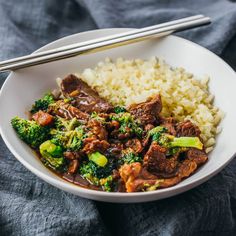 a white bowl filled with beef and broccoli on top of rice next to chopsticks