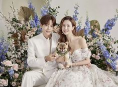 a man and woman sitting next to each other holding a teddy bear in front of flowers