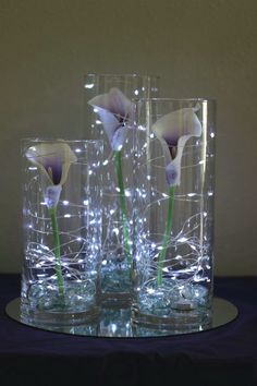 three glass vases sitting on top of a table with flowers in them and lights behind them