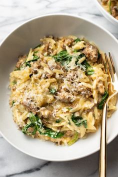 a white bowl filled with pasta and meat on top of a marble table next to two gold forks