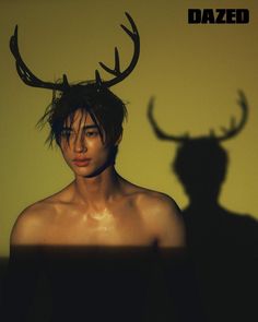 a shirtless man with long hair and antlers on his head, standing in front of a yellow wall