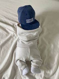 a baby wearing a baseball cap and sweatpants laying on a bed with white sheets