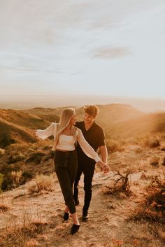 a man and woman walking down a dirt path in the desert at sunset with mountains in the background