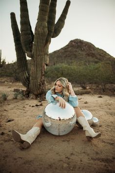 a woman sitting on the ground in front of a cactus and holding a large round object