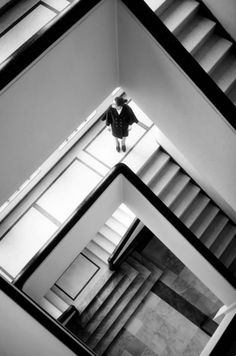 black and white photograph of person standing on top of stairs in an office building, looking up at the ceiling