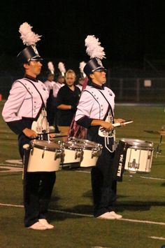 two men in marching uniforms playing drums on a field with other people standing behind them