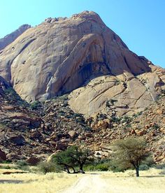 a dirt road in front of a large rock formation with trees and bushes on the side