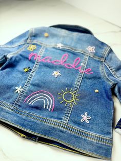 a denim jacket with embroidered words and rainbows on the back, sitting on a white surface