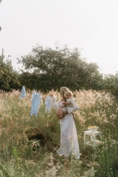 a woman in a white dress holding a baby standing in a field full of clothes