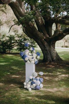 a tall white and blue floral arrangement on the ground in front of a large tree