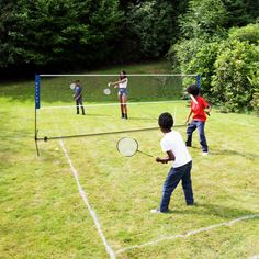 four people playing tennis on the grass in front of a fenced area with trees