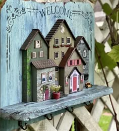 a wooden sign that says welcome with small houses on it and potted plants in the window